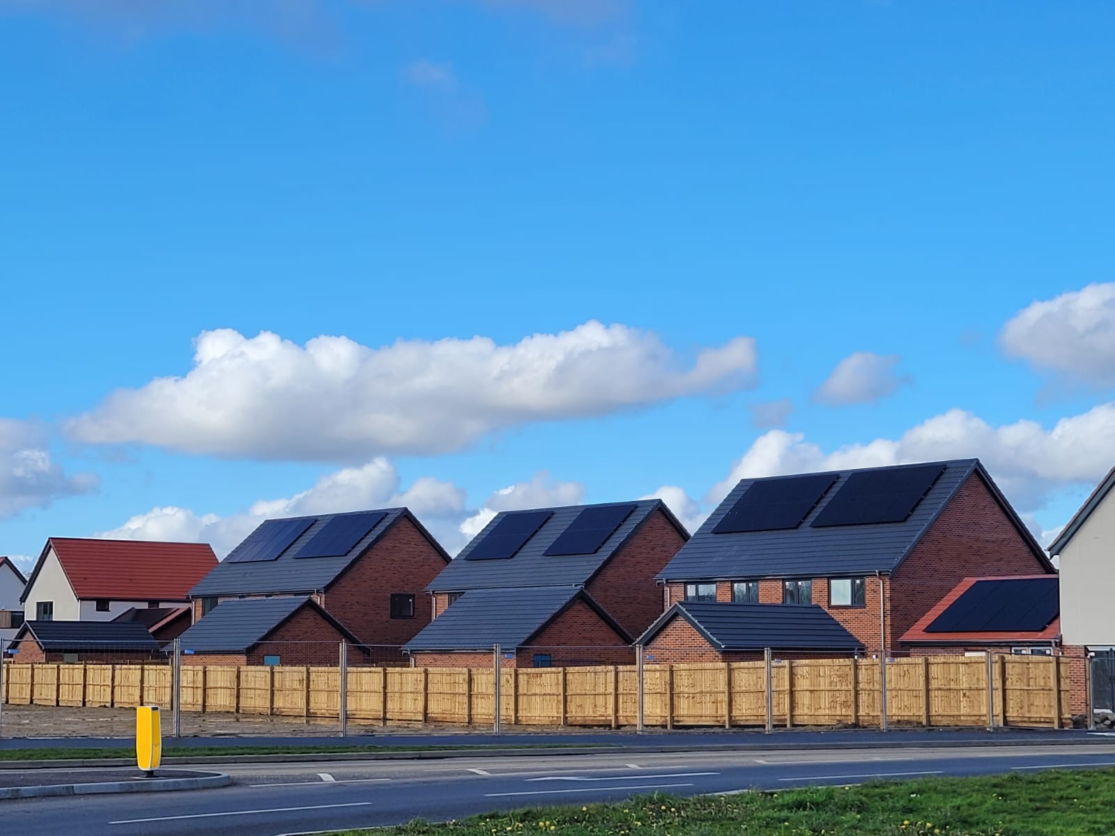 A row of modern houses with solar panels on their roofs, bordered by a wooden fence, under a blue sky with scattered clouds.