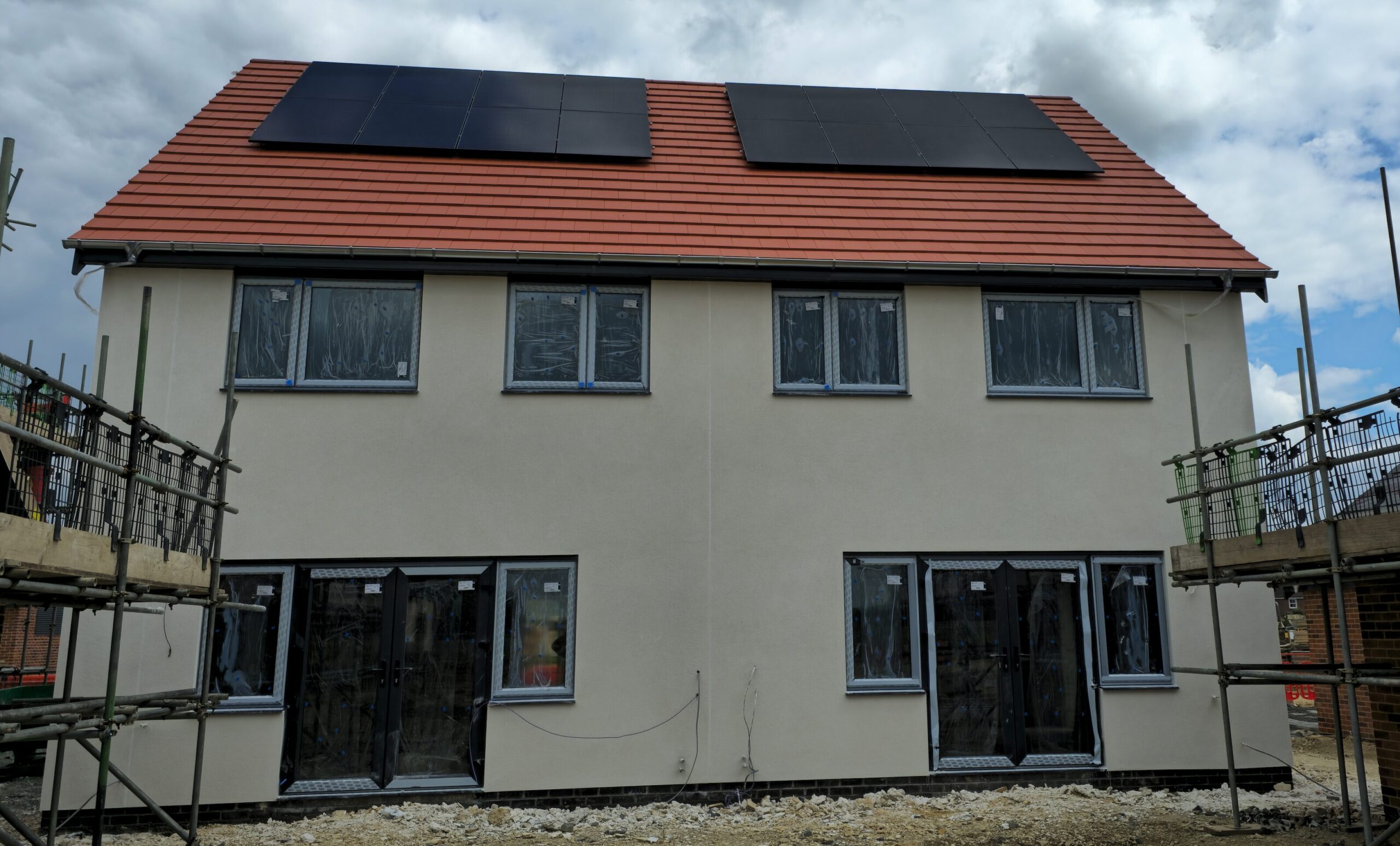 A newly constructed two-story house with solar panels on the roof, scaffolding on both sides, and windows covered in protective film.