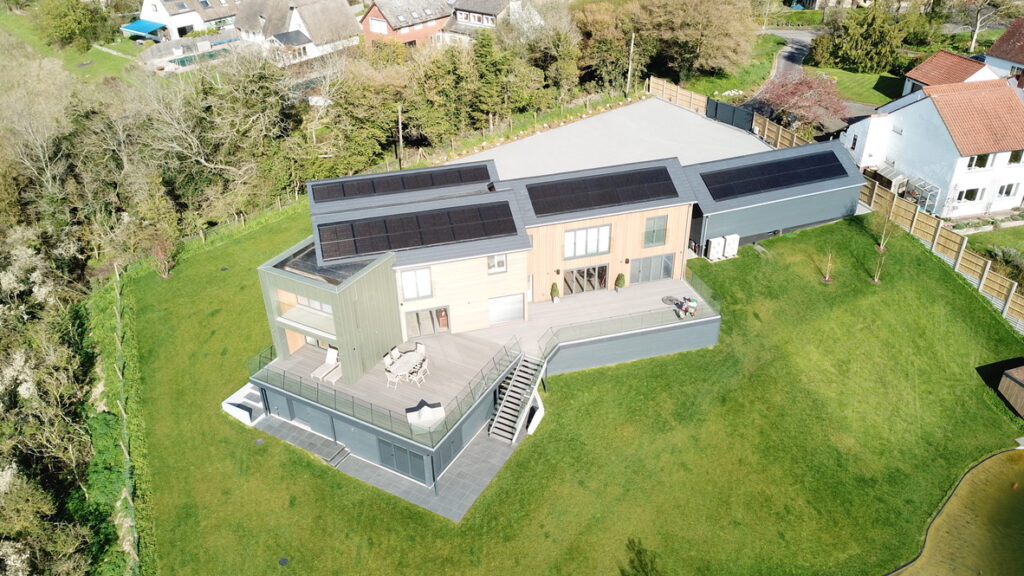 Aerial view of a modern, multi-level house with solar panels on the roof, surrounded by a spacious green lawn and trees.