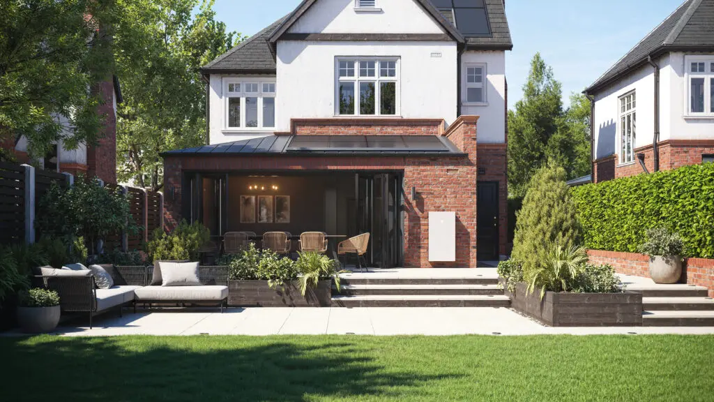 The garden of a modern house, with brickwork. A Tesla Powerwall is affixed to a wall.