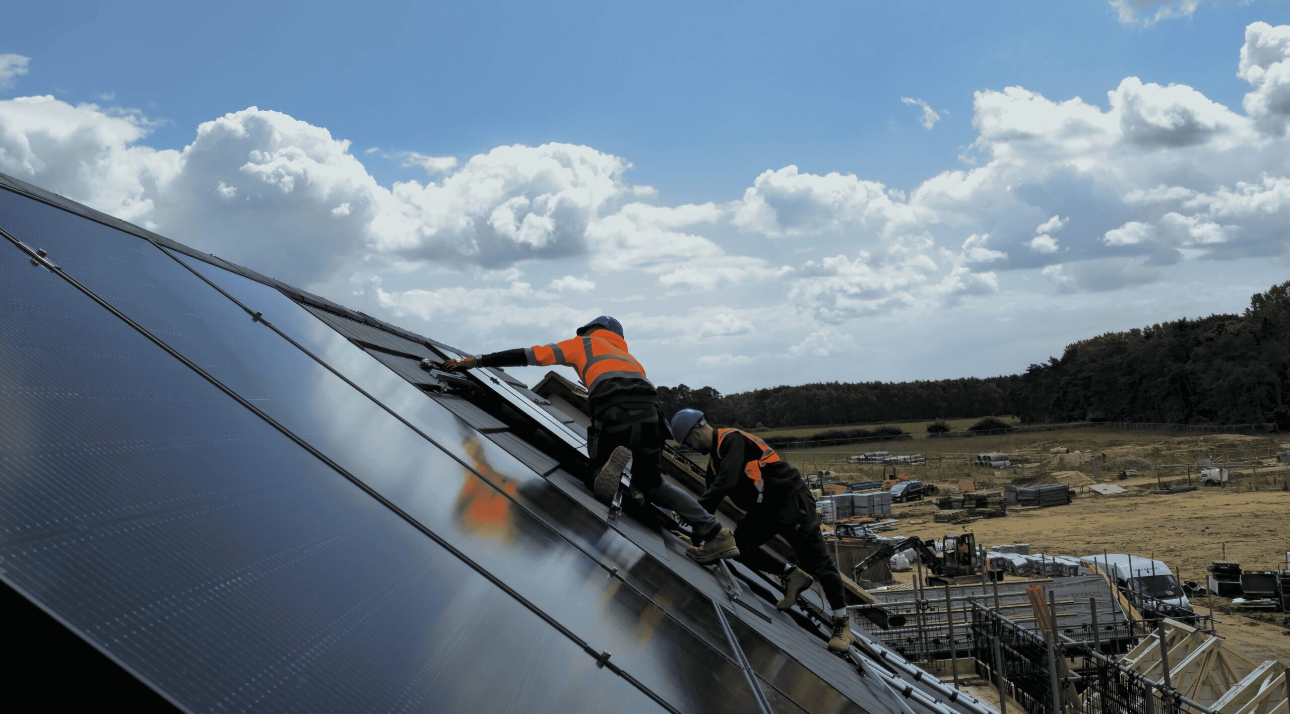 Two workers in safety gear install solar panels on a sloped rooftop under a partly cloudy sky, with construction equipment and a field visible in the background.