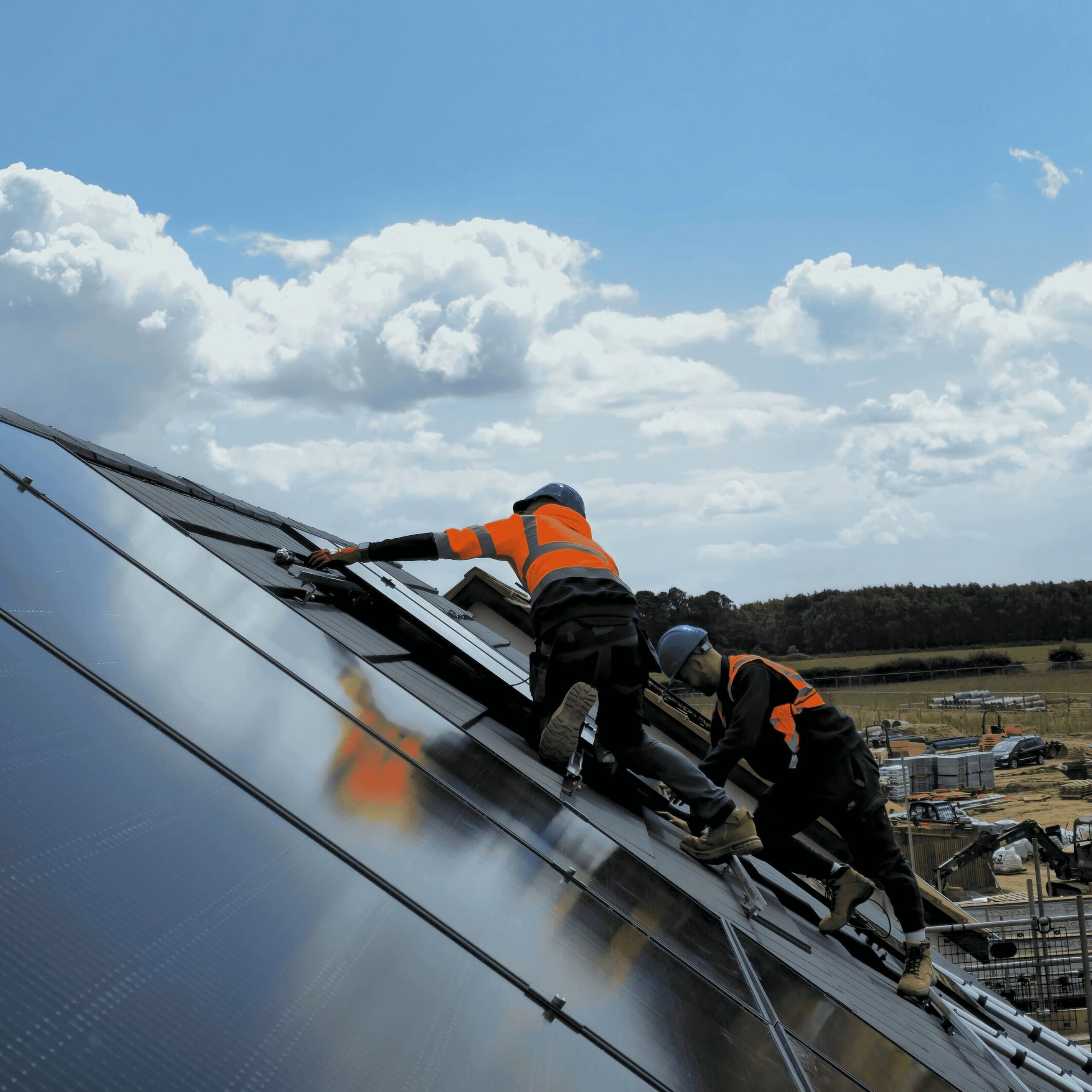 Two workers in safety gear install solar panels on a sloped roof under a partly cloudy sky, with a rural landscape visible in the background.