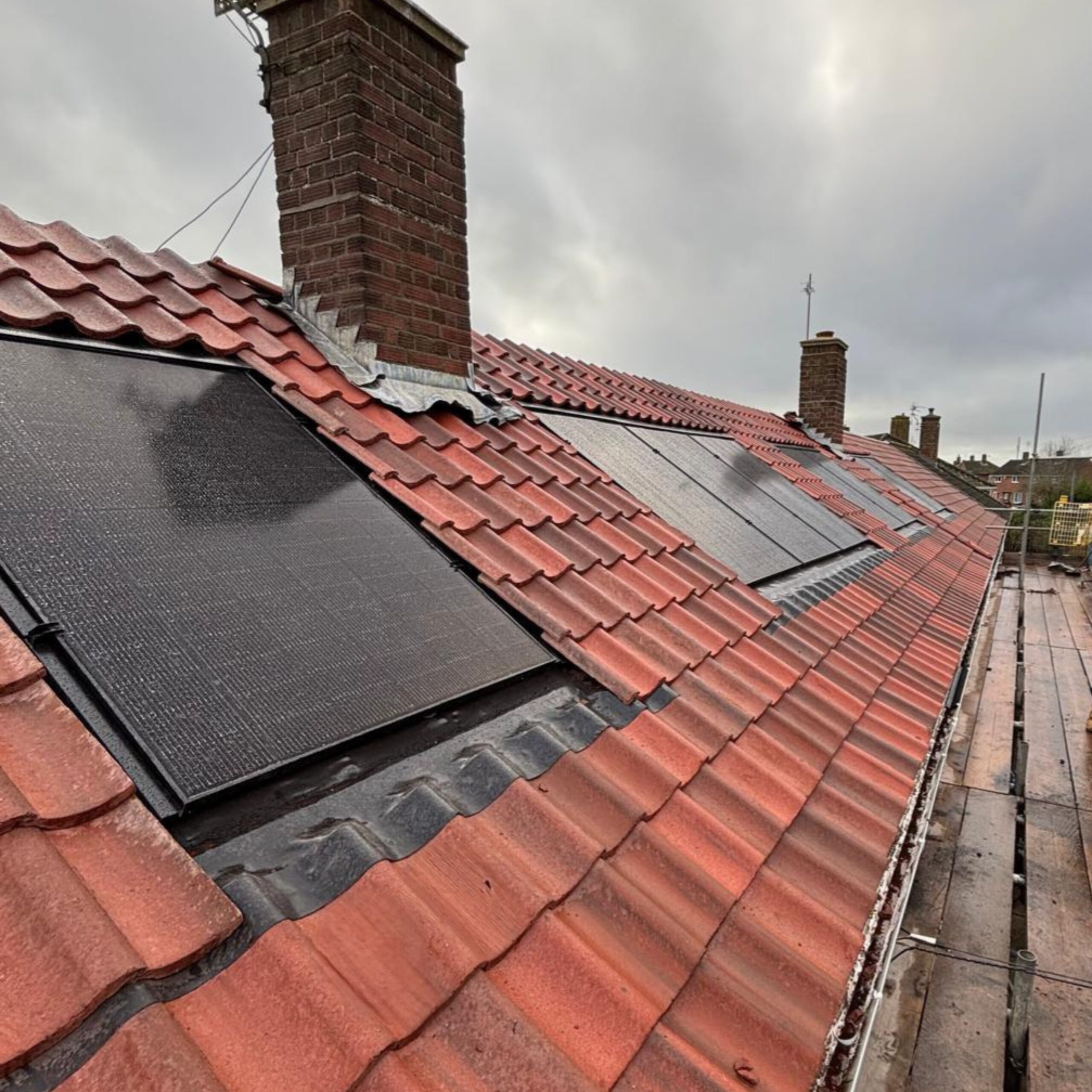 Solar panels are installed flush with the red tiled roof of a residential building under a cloudy sky, with brick chimneys visible.
