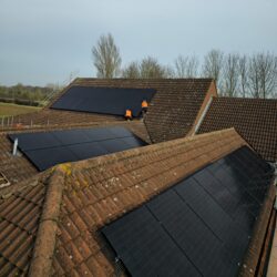 Two workers in orange vests install black solar panels on the tiled roofs of residential buildings, with trees and a field in the background.