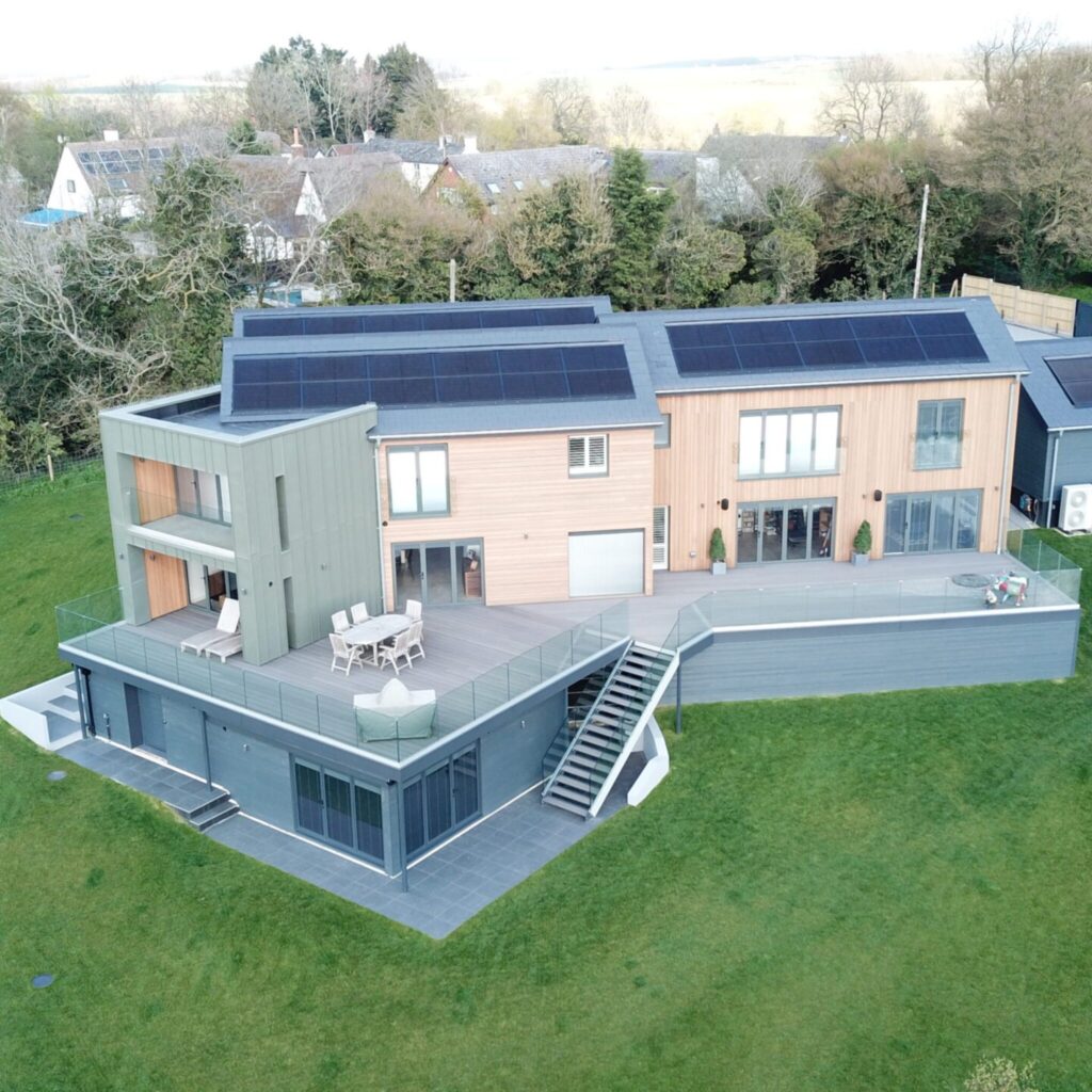 Modern two-story house with large deck, glass railings, solar panels on the roof, and surrounded by grass lawn.