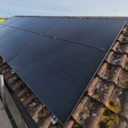 Eight black solar panels installed on a sloped, mossy tiled roof with scaffolding set up alongside the roof edge.