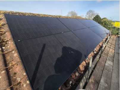 Six black solar panels are installed on a sloping roof next to a wooden platform, with trees and a clear sky in the background.
