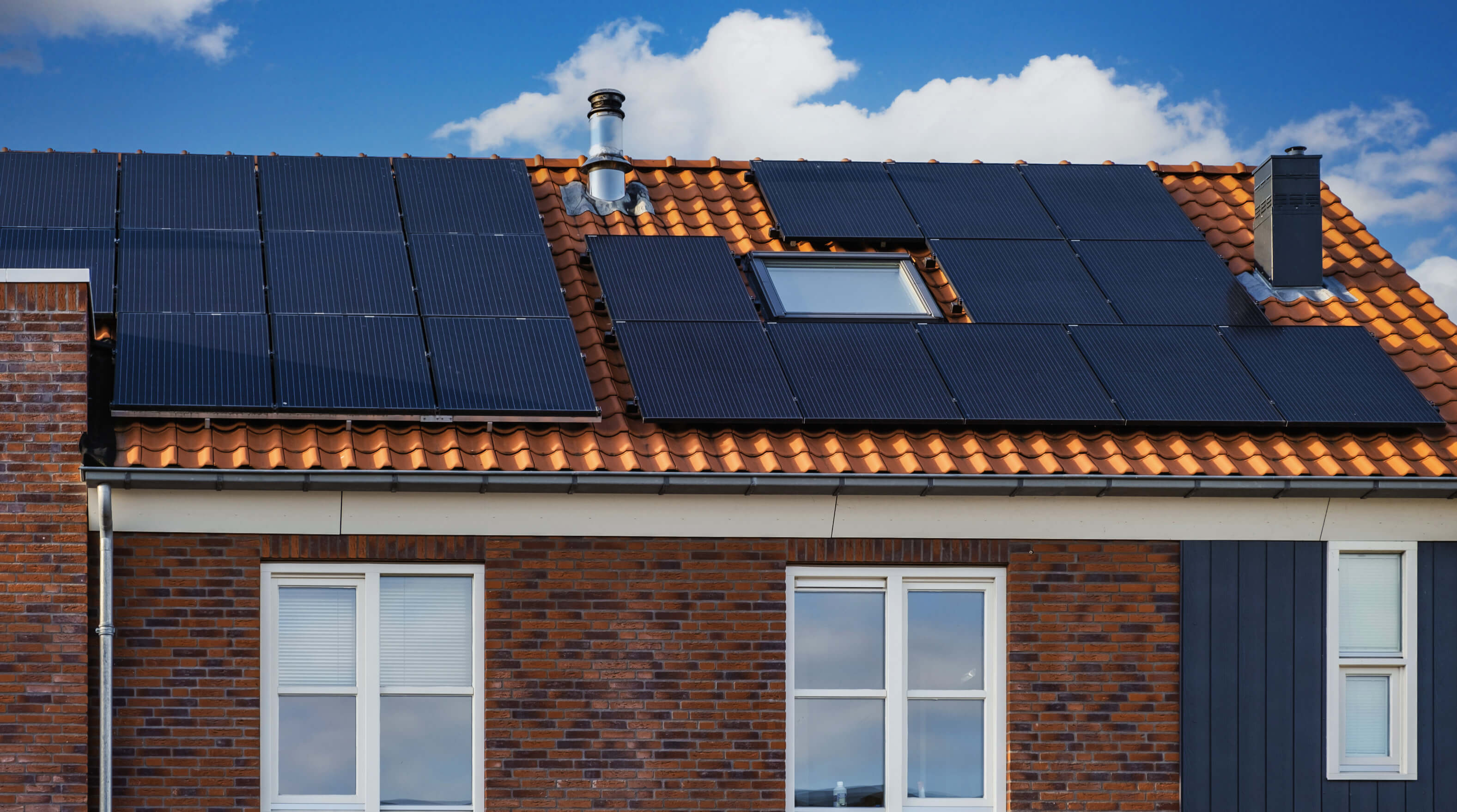 A brick house with a tiled roof has multiple black solar panels installed around a skylight, under a blue sky with clouds.